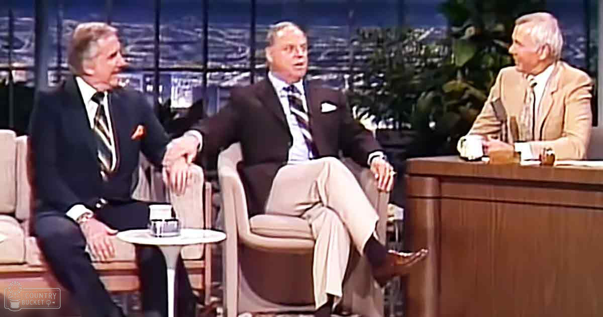 An indoor talk show set with three older adult males wearing classic dark suits and patterned ties against large windows showing a cityscape at night.