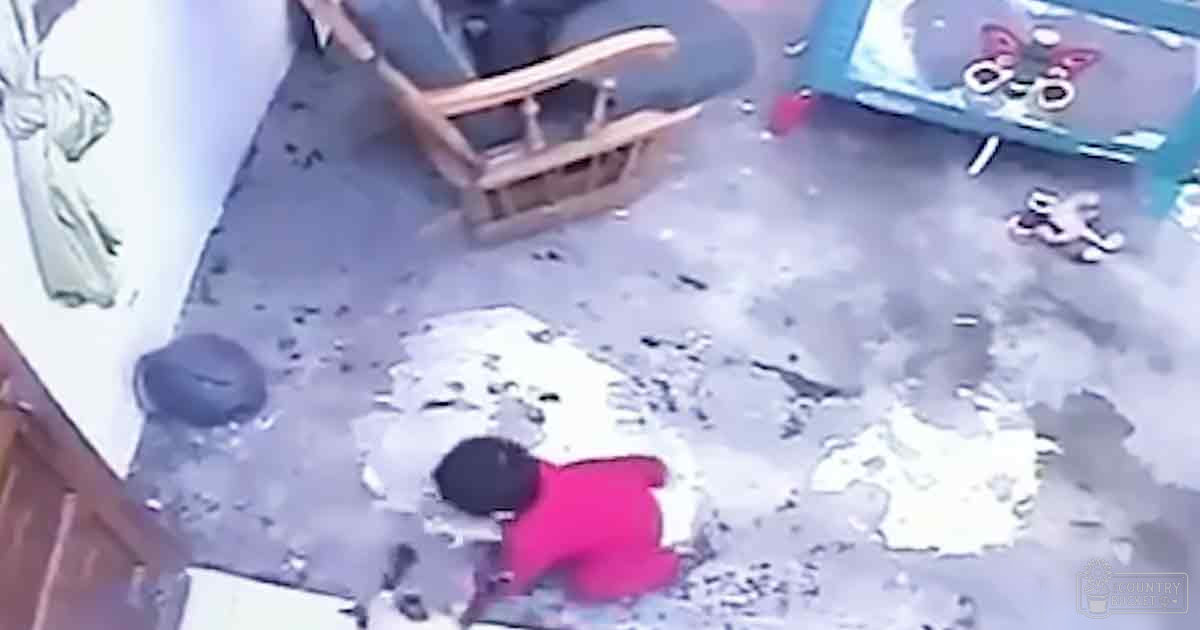 A toddler in red shirt, dark pants, and possible black hat or hood plays with toys scattered around a room containing a wooden rocking chair and patterned rug.