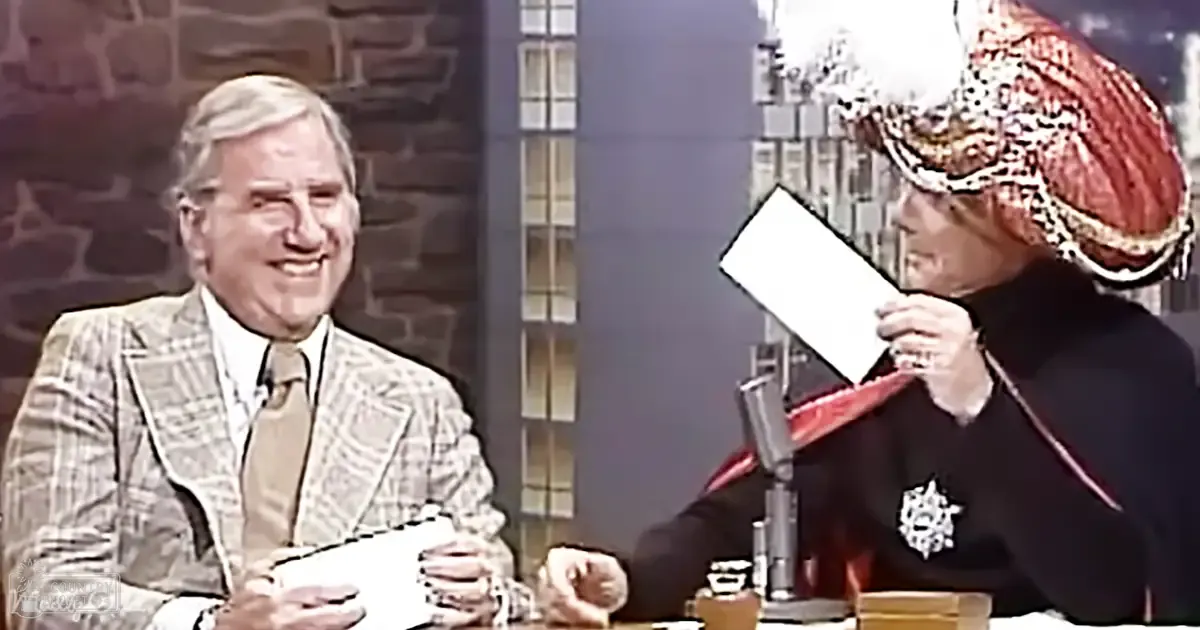 A middle-aged male talk show host in a plaid blazer and white shirt, seated with an older adult female guest wearing a black top and red/gold patterned headscarf, in a formal indoor studio setting with brick walls and large windows.