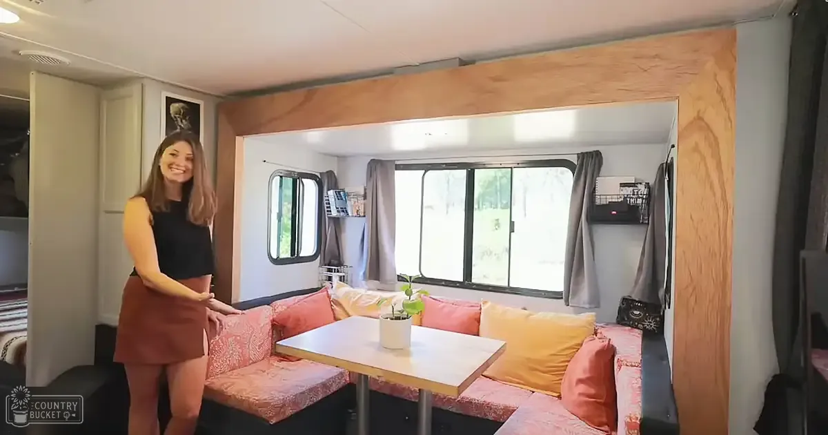 A cozy living space within an RV interior with a sofa with pink and yellow cushions, a small white table holding a plant, curtains over the windows, shelves with books and decorative objects.