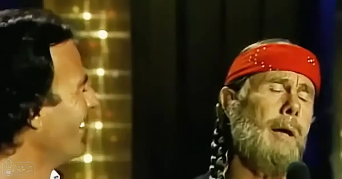 A man with a red headband and braided hair stands on stage holding a microphone during an awards ceremony or performance, while another adult male singer in dark attire with a patterned shoulder design stands nearby.