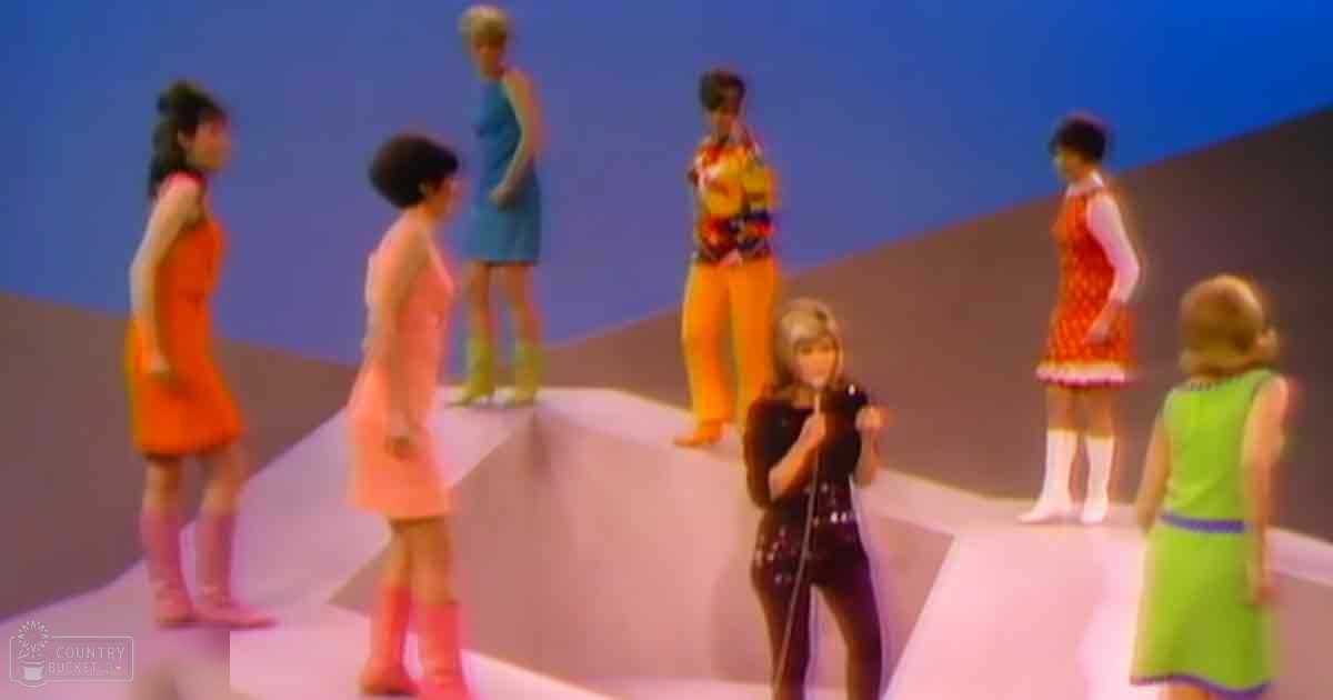 A young adult female performer singing into a microphone with children and adults around her on an indoor stage setup with a simple blue and gray backdrop.