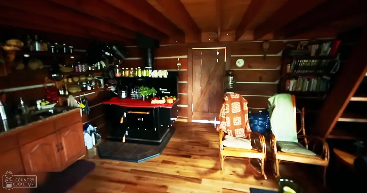 Cozy cabin interior with wooden furniture, produce on countertop, patterned chairs, books on shelves, rustic setting, evening lighting.