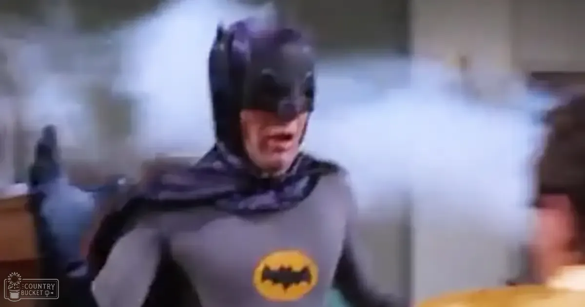 A young adult male dressed as Batman stands indoors, possibly in a residential kitchen with artificial lighting and dark gray and yellow tones.