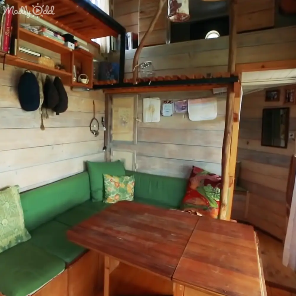 A cozy cabin interior with a wooden dining table, green cushioned seating, decorative pillows, shelves holding books and cups, and a hanging rack for hats. The setting suggests daytime natural lighting in an indoor rustic environment.