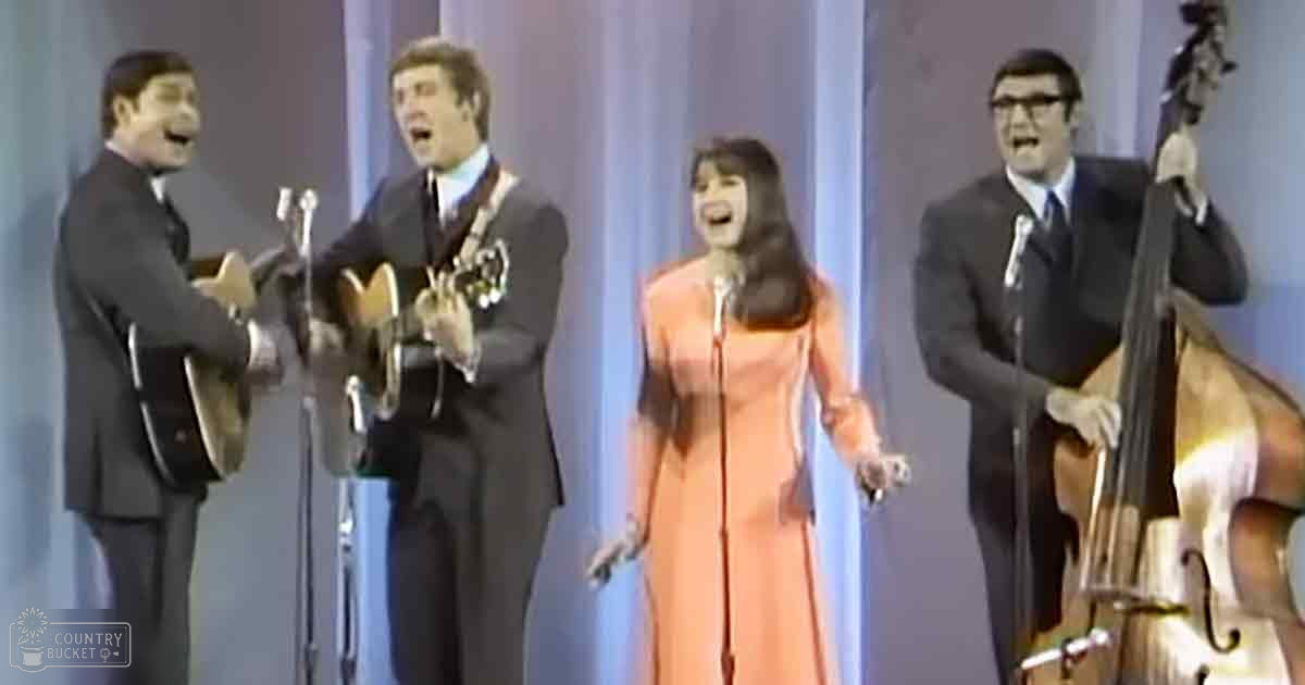 A musical performance with four adults singing and playing acoustic guitars against a simple blue curtain backdrop under bright stage lighting.