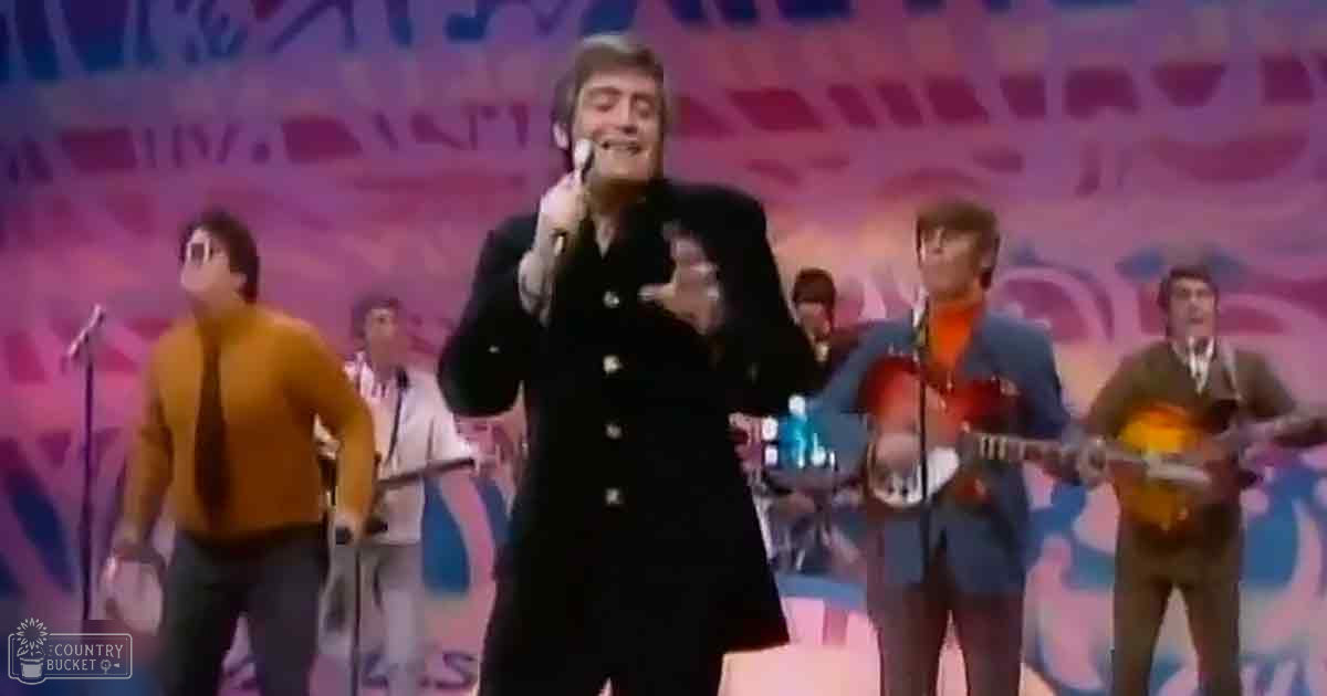 Adult male singer wearing a black double-breasted suit jacket and dark pants, holding a microphone; other performers include an adult male in an orange sweater and another in a white outfit on stage with colorful abstract backdrop.