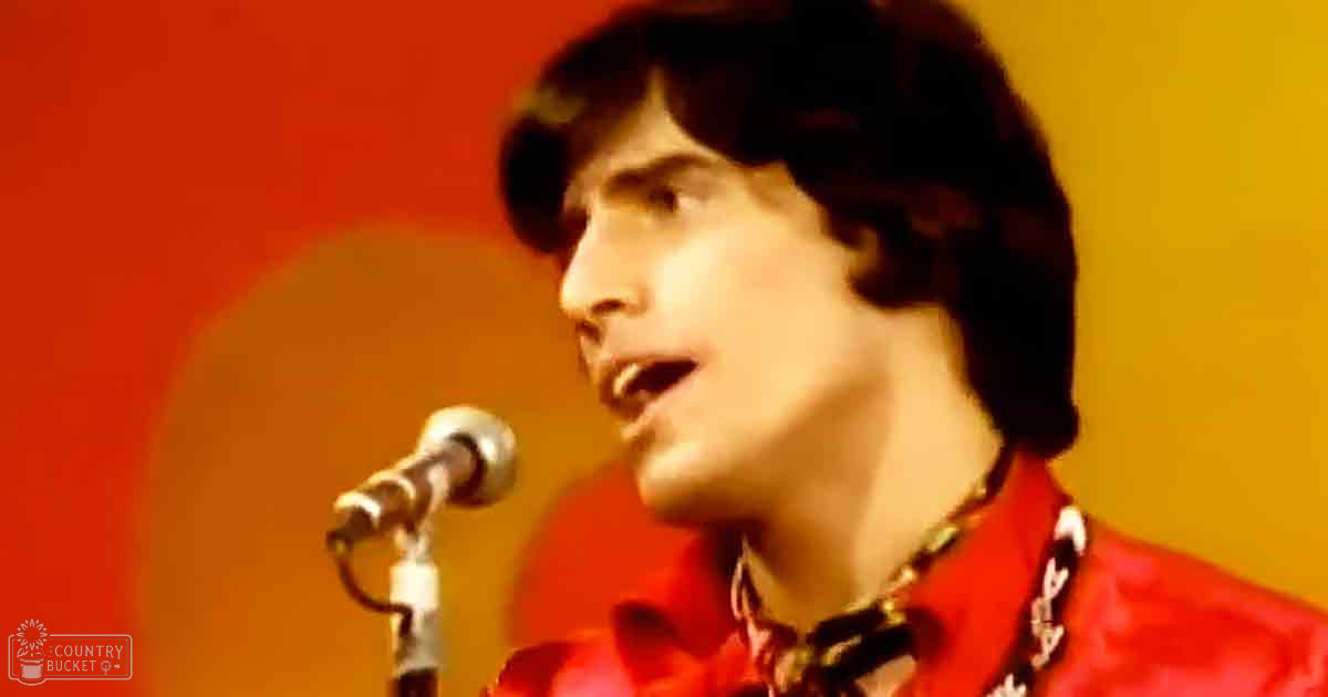 A young adult male singer in a red jacket with white and black patterned lapels performs at an indoor event, wearing a gold chain necklace.
