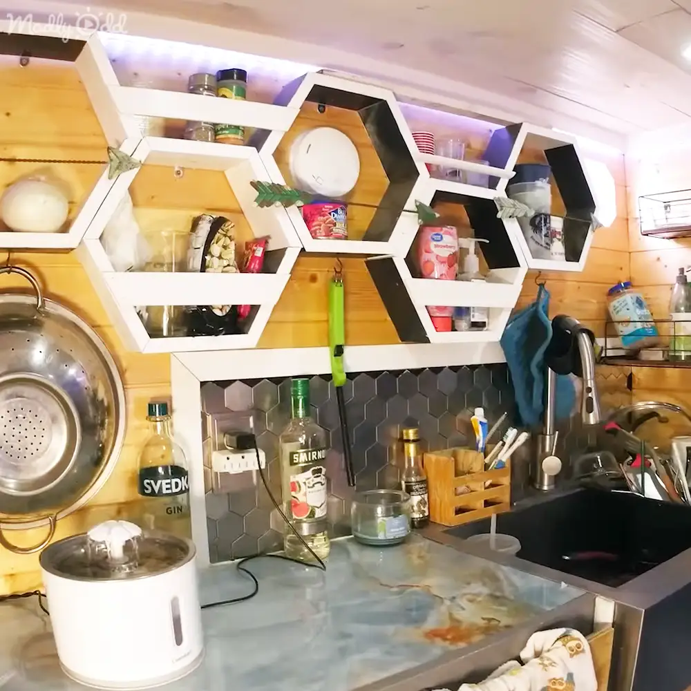 A cozy kitchen setup with hexagonal shelving unit, colander, bottles of Svedka vodka and Smirnoff gin, wooden crate holding toiletries, sink, and a white appliance in a daytime scene with natural light streaming from the right side. The setting includes light wood tones on walls, white shelves and appliances, black hexagonal shelving unit, and various colored items stored.