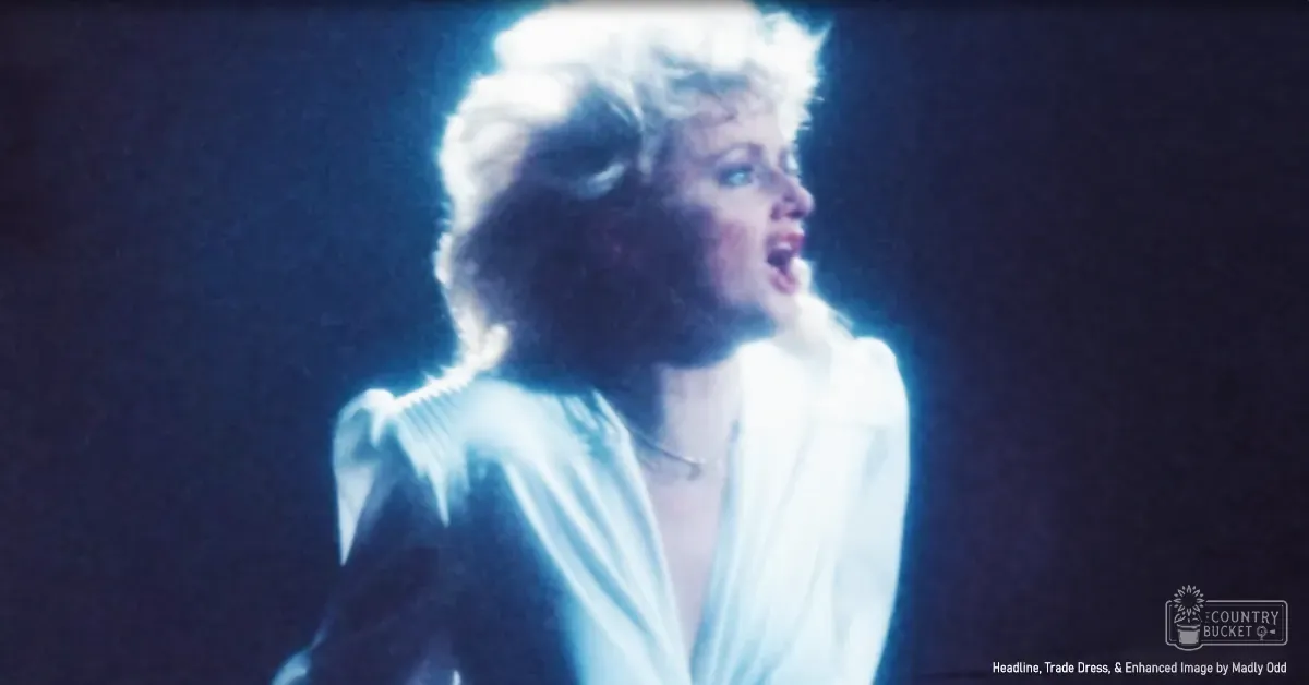 An adult female performer in a stage setting, wearing a white formal dress with wide shoulder straps from an earlier era under dramatic stage lighting.