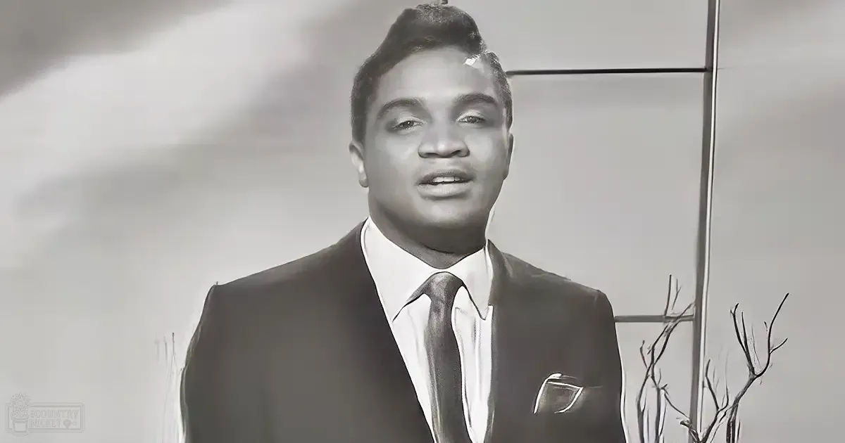 A middle-aged adult male wearing a dark, fitted suit jacket with a pocket square and a collared shirt, seated in a modern indoor setting with decorative elements like branches and abstract art on the wall.