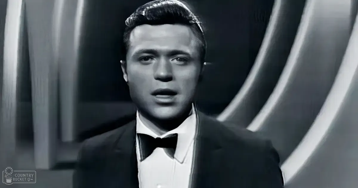 Young adult male in a formal tuxedo stands indoors against a dark, curved architectural backdrop.