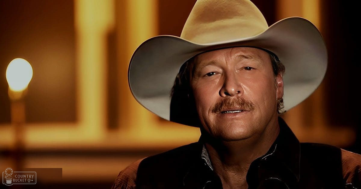 A middle-aged adult male in a relaxed fit dark brown and black cowboy shirt with long sleeves, wearing a wide-brimmed cowboy hat with light-colored fabric and an earring in his left ear, set against warm-toned indoor lighting suggesting an interview or photography session.