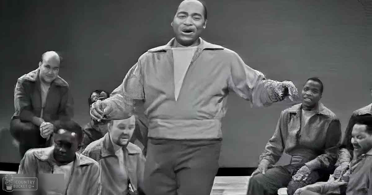 Adult male singer in a mid-20th-century style jacket on stage with seated individuals, indoor performance venue, muted grayscale lighting
