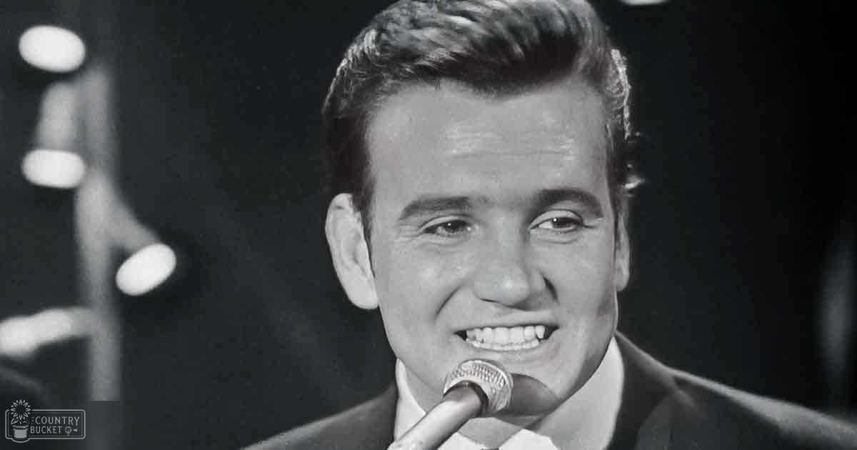 A young adult male in a tailored suit jacket, white shirt, and tie stands on stage with a microphone, performing under focused indoor lighting.