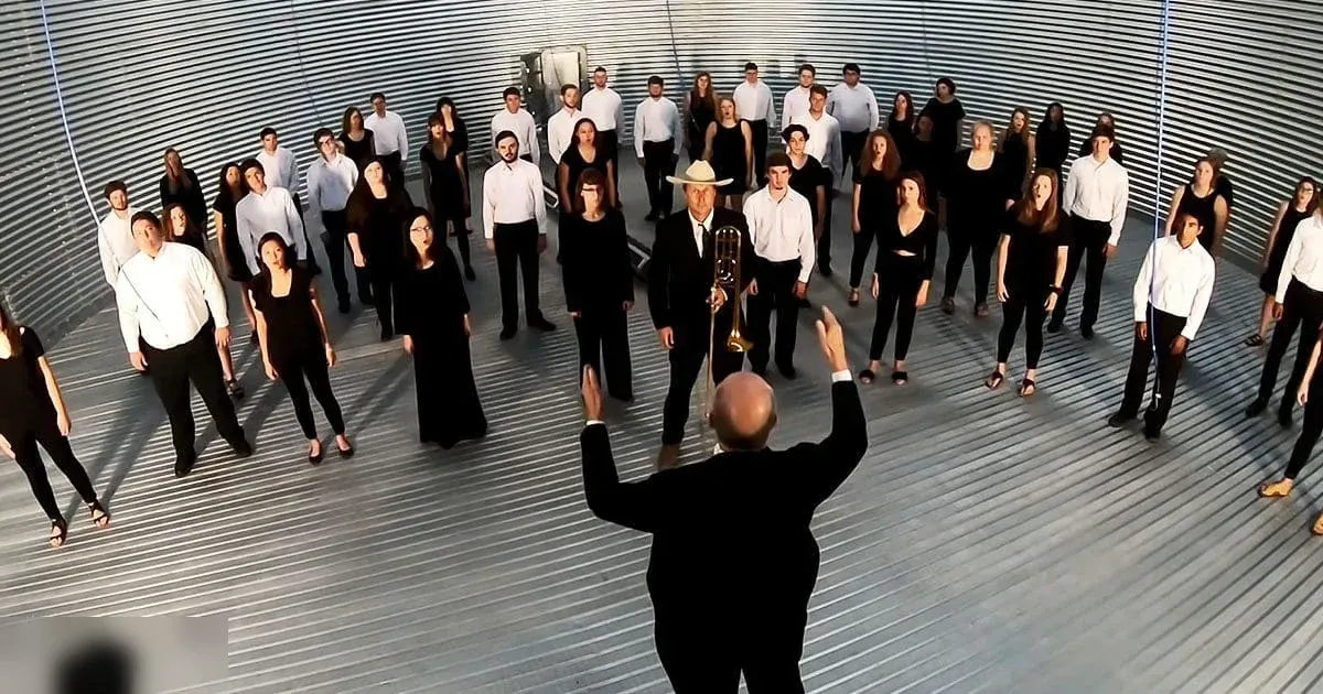 Adult male conductor leading a choir in an indoor performance setting with metallic walls, natural daylight, and choir members dressed in black or white attire.