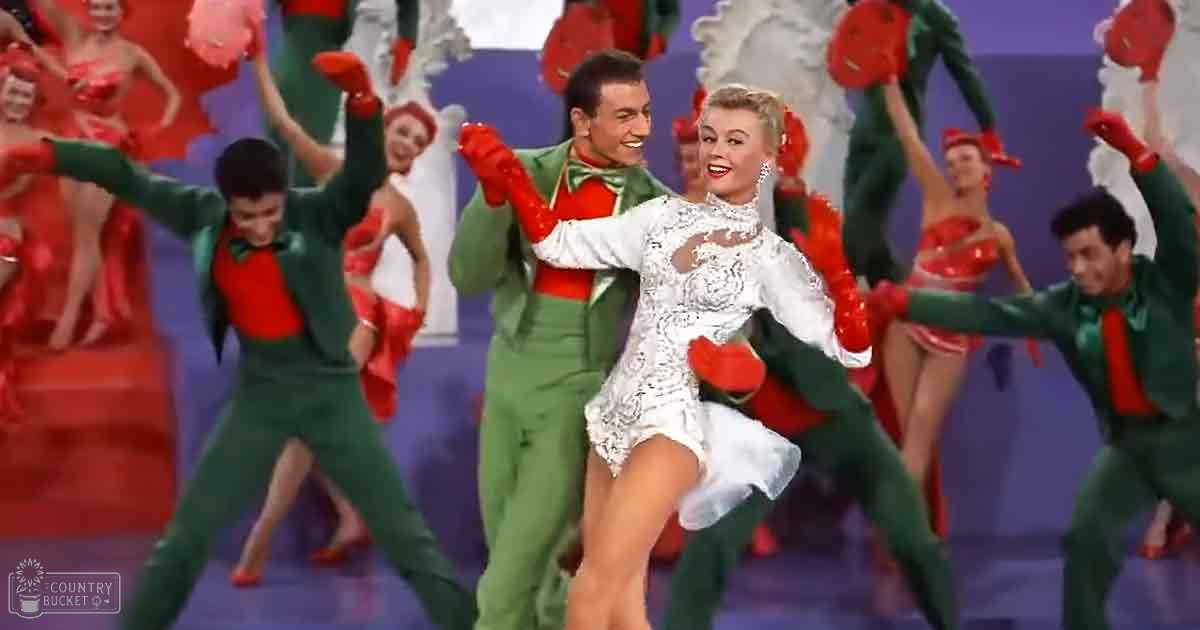 A young adult female dancing in a festive setting, wearing a white lace dress with green gloves and red shoes. Background dancers are dressed in green pantsuits with red shirts and black bow ties.