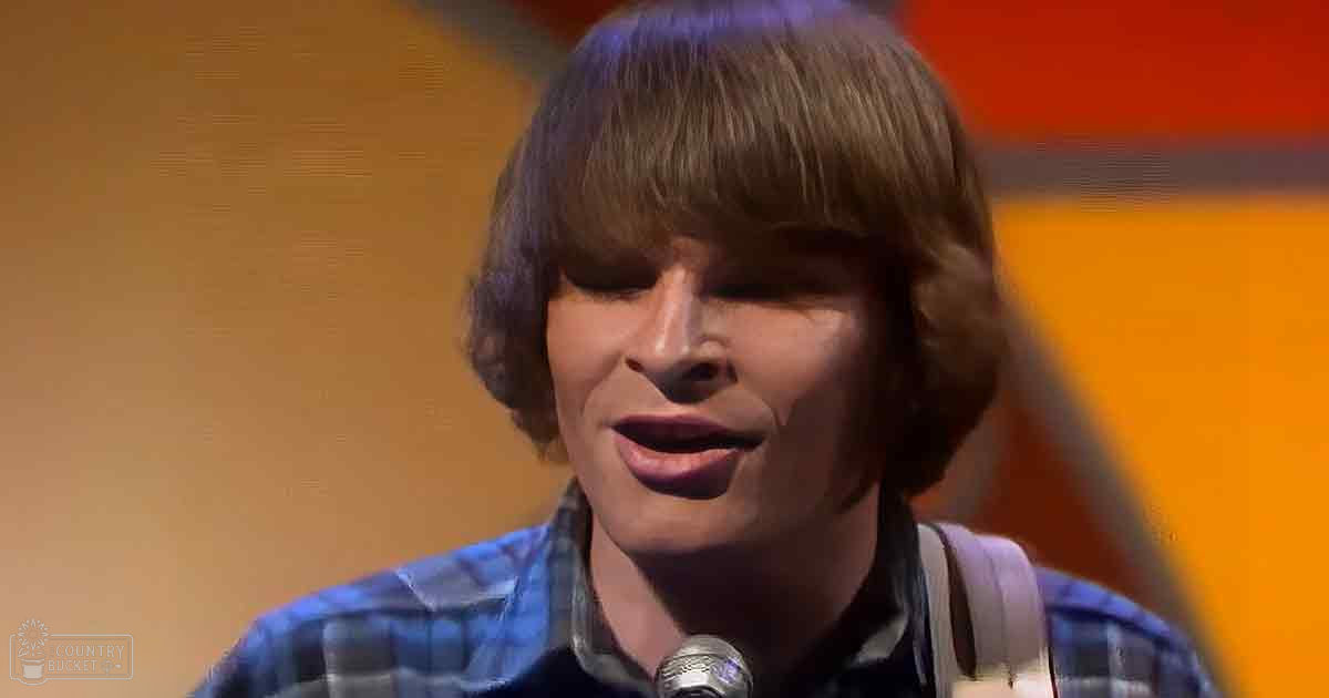 A young adult male in a relaxed fit, button-down style plaid shirt with long sleeves, holding a microphone close to his mouth, likely at a performance venue with warm lighting and orange-yellow tones.