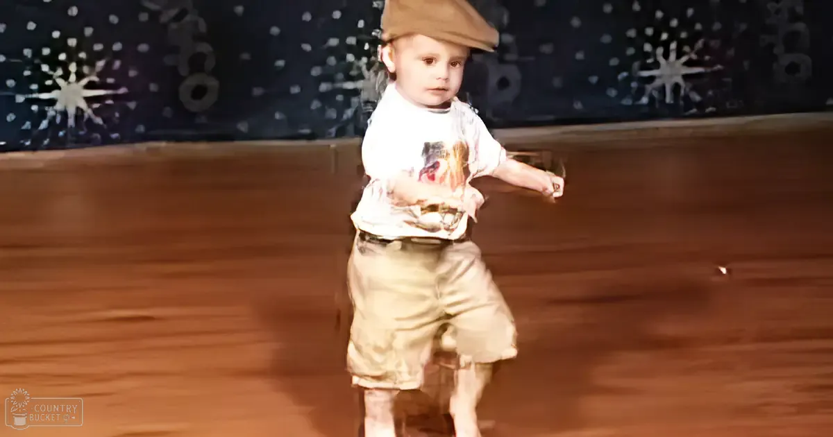 A toddler wearing a white graphic t-shirt with a red element, khaki shorts, and a brown beret-style hat stands indoors in what appears to be a performance setting with decorative snowflake or star backdrop suggesting a winter theme.