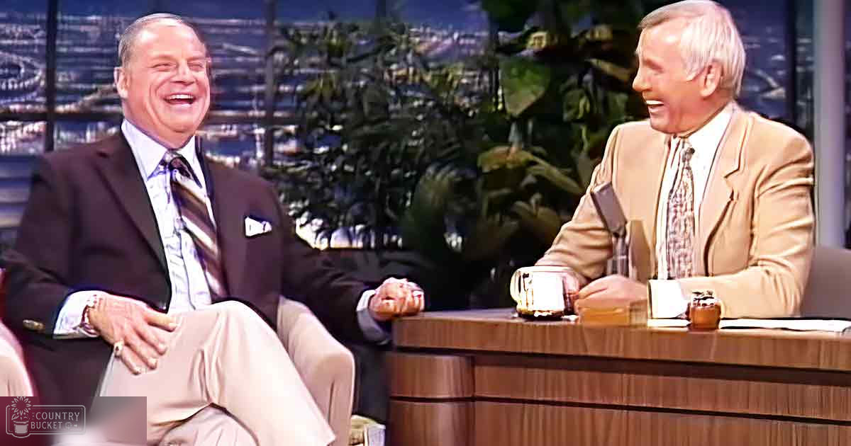 A middle-aged man in a dark suit jacket with a patterned tie sits beside an older adult male wearing a light beige suit jacket and white shirt, both on a talk show set with a cityscape backdrop at night or dusk.