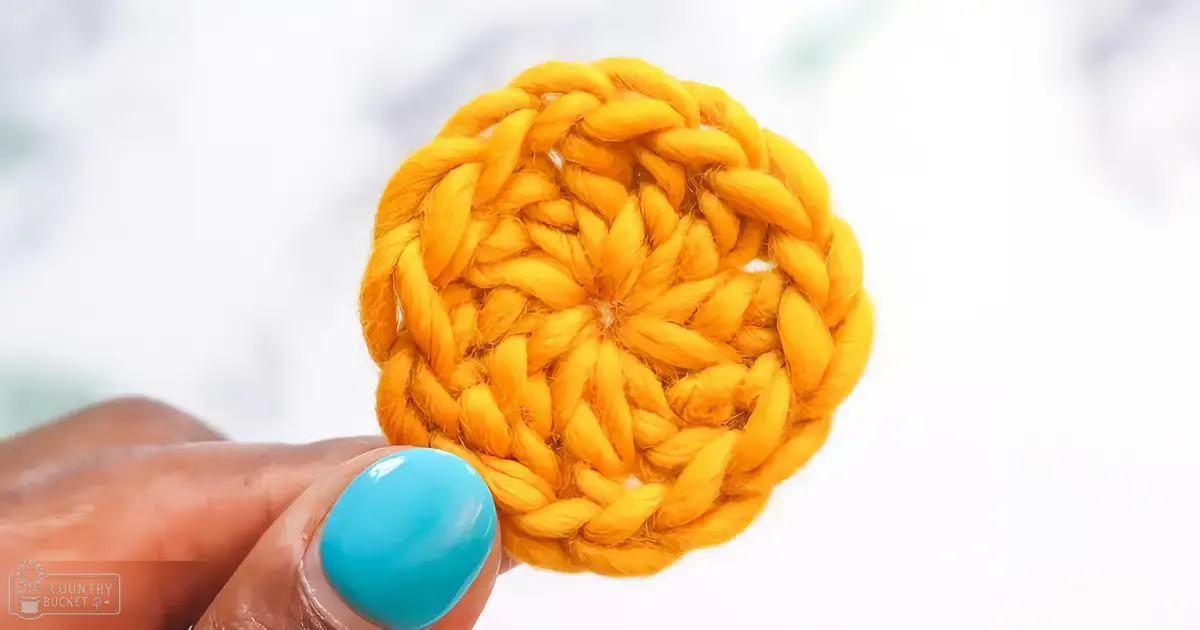 A close-up of a hand holding a yellow crocheted circle with blue nail polish, set against a light-colored surface indoors.