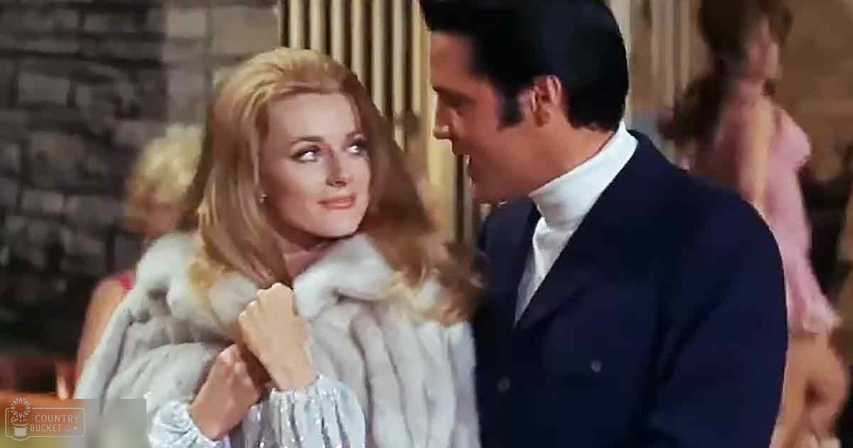 A couple in an indoor setting, woman wearing a luxurious white fur coat and matching gloves with light blue dress underneath; man dressed in a dark navy suit jacket over a white turtleneck sweater.