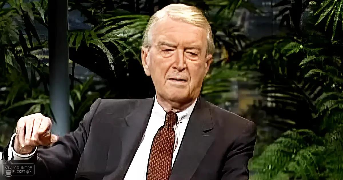 An older adult male in a formal suit and tie, seated against a backdrop of greenery suggesting an indoor setting with natural light.