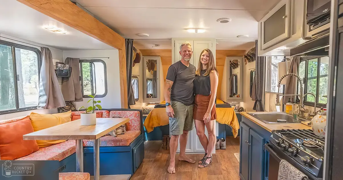A couple inside an RV interior with wooden dining table and blue cabinets during the daytime.