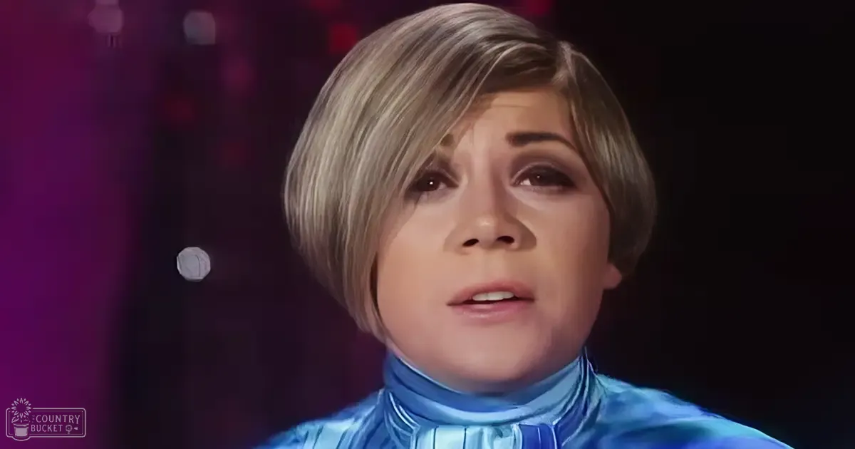 A young adult female wearing a shiny blue, high-collared garment with vertical stripes stands indoors under stage lighting. A small white circular object floats near her head on the left side of the frame.
