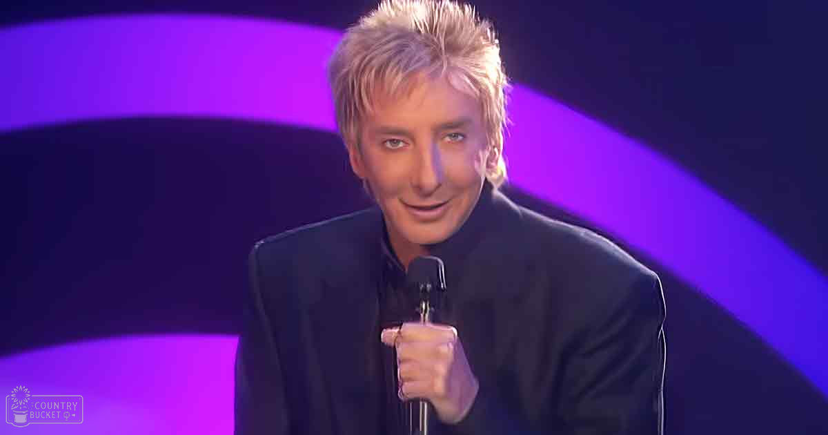 An adult male performer in a dark suit jacket with high collar and long sleeves, holding a microphone on stage under purple lighting at an indoor performance venue.