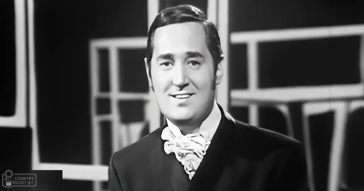 A young adult male wearing a tailored black suit with a patterned bow tie, standing indoors against a geometric wall backdrop.