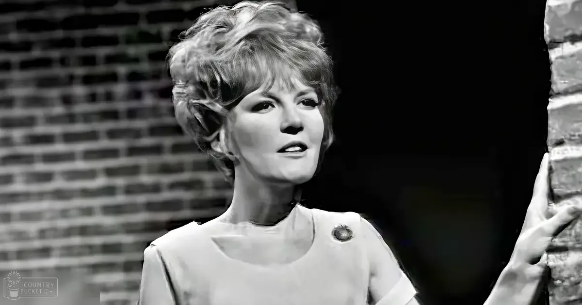 A middle-aged woman in a sleeveless, knee-length dress with a round neckline, standing against brick walls in an urban setting at dusk.