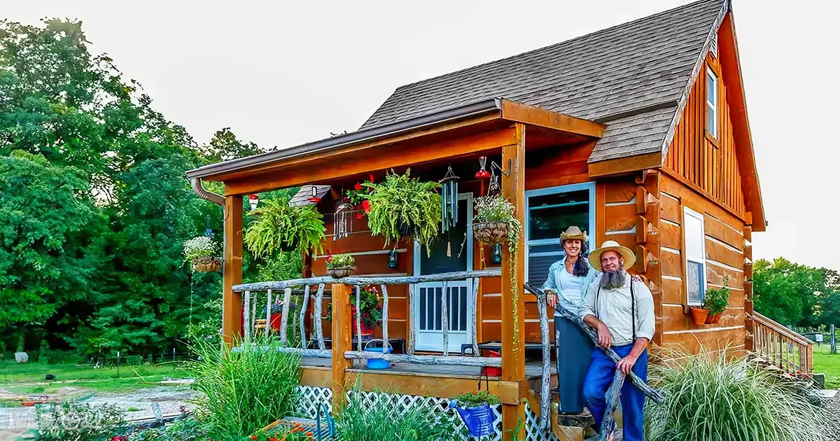 A middle-aged adult female in a white shirt and blue jeans stands with an older male holding a wooden tool or stick near their rustic cabin home surrounded by lush greenery, suggesting spring or summer.