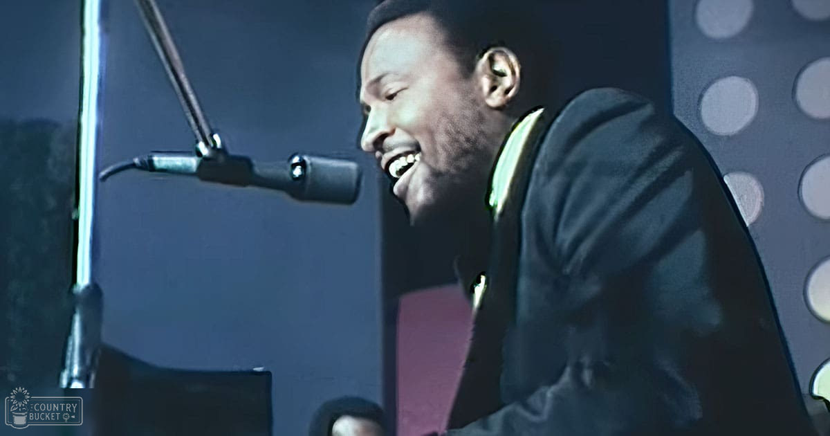 A male adult singer wearing a dark-colored, slightly tailored jacket with visible buttons and light green collar in a recording studio setting.