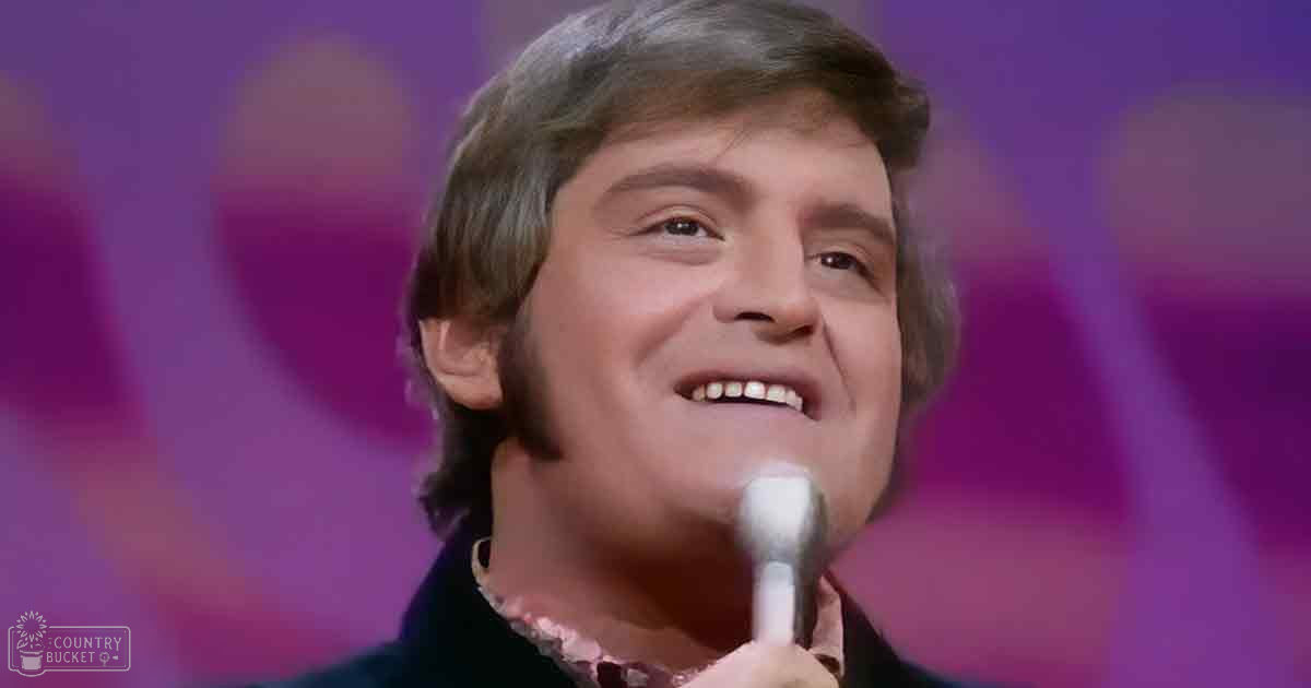 A middle-aged adult male singer wearing a dark jacket with a high collar, patterned shirt, and singing into a microphone in an indoor performance setting.