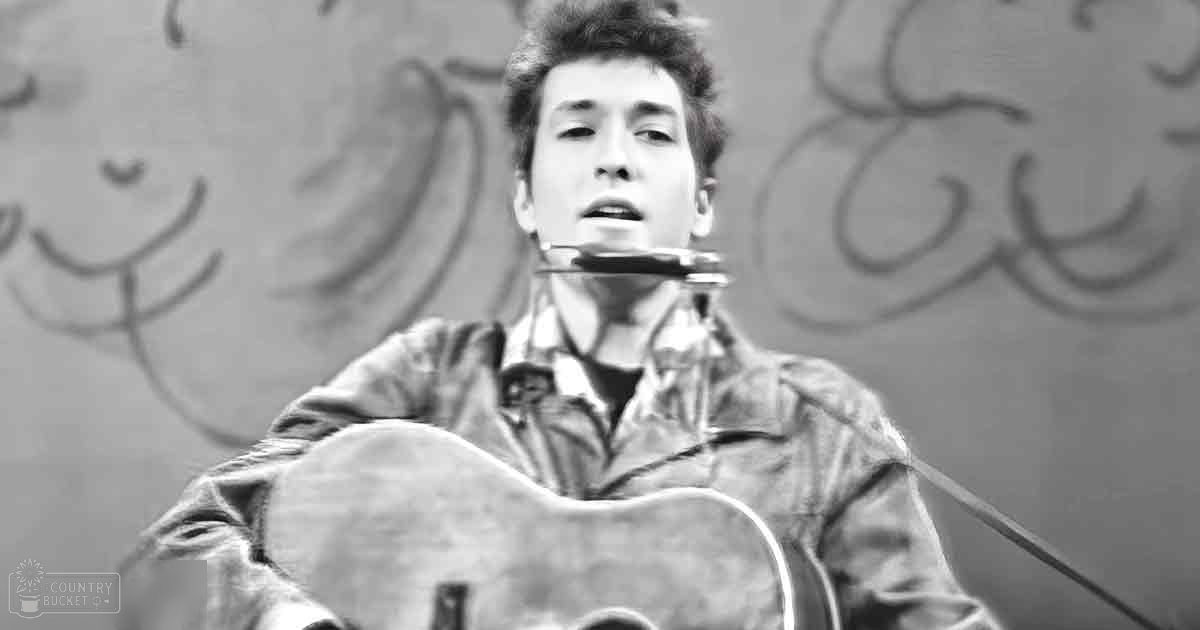 A young adult male wearing a relaxed fit jacket with a collar, buttoned up shirt, playing an acoustic guitar on what appears to be an urban stage-like setting.