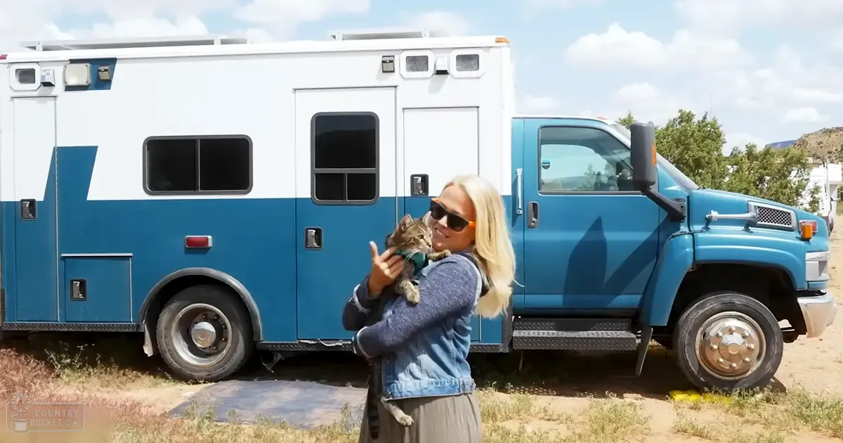 An adult female holds a tabby cat in front of an RV amidst dry grass and trees under bright daylight.