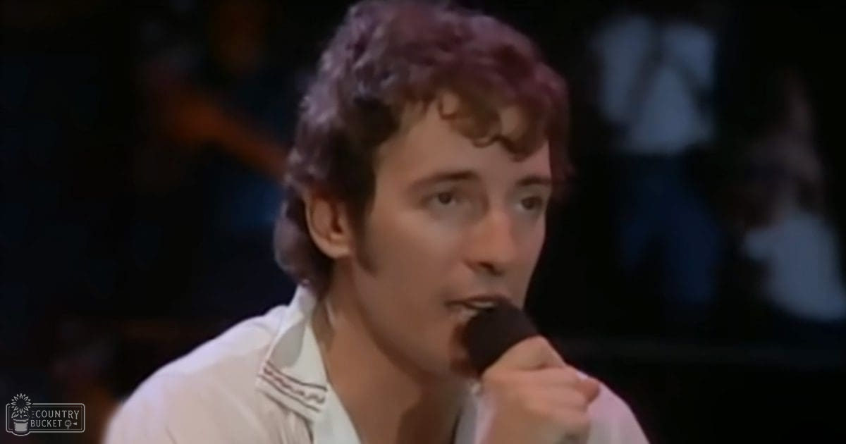 A young adult male singer performing at a concert, wearing a white shirt with striped shoulders, against a dark indoor performance backdrop.