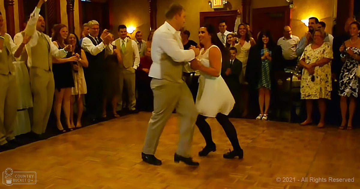 A couple dancing at an indoor wedding reception, surrounded by guests in formal attire under warm artificial lighting.