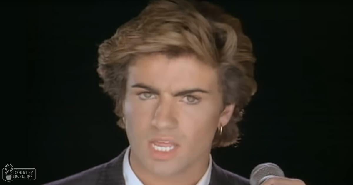 A young adult male in a dark suit jacket over a light-colored collared shirt stands against a black backdrop with a microphone visible, suggesting a performance or interview setting from the late 1980s to early 1990s.