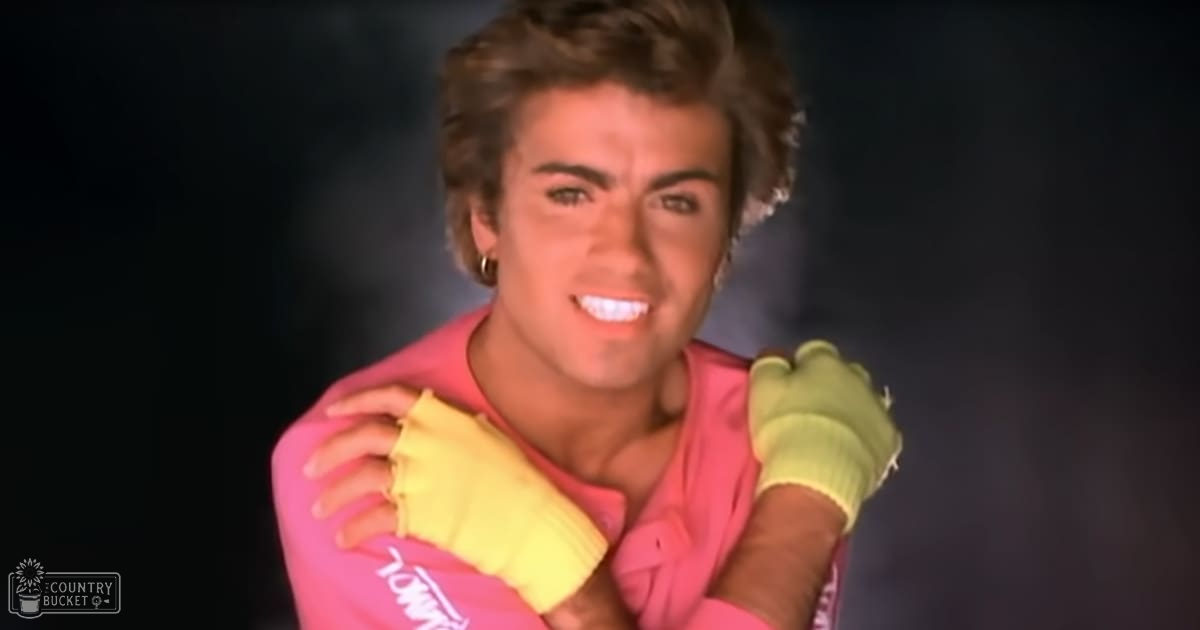 A young adult male in a casual, loose-fitting, bright pink sleeveless top with gold hoop earrings, set against a dark studio background.