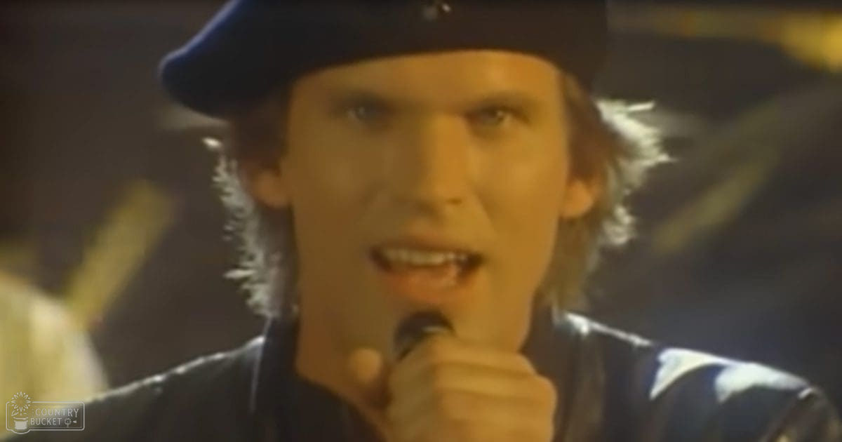 A young adult male singing into a microphone in what appears to be an indoor performance setting, wearing a dark-colored jacket with light patterns and a black hat.