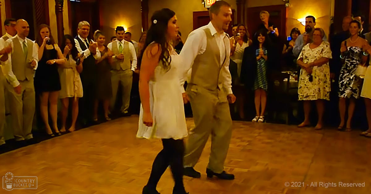 A couple dancing at an indoor wedding reception with guests in formal attire, warm indoor lighting and wooden flooring.