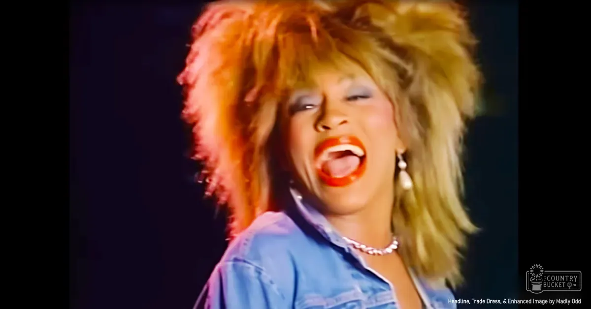 A middle-aged female performer wearing a denim shirt with pearl earrings and necklace in an indoor or stage setting, likely for a music video.