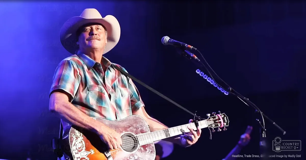 A middle-aged male performer wearing a white cowboy hat and a short-sleeved button-up shirt with tie-dye patterns at a concert setting under blue stage lighting.