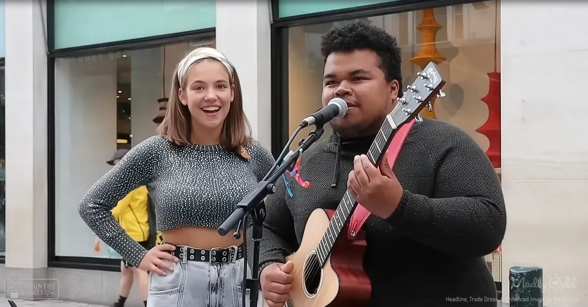 A young adult female and an adult male performing street music, wearing a long-sleeved crop top with white polka dots and a textured sweater or jacket. Urban setting suggests near a shopping area.