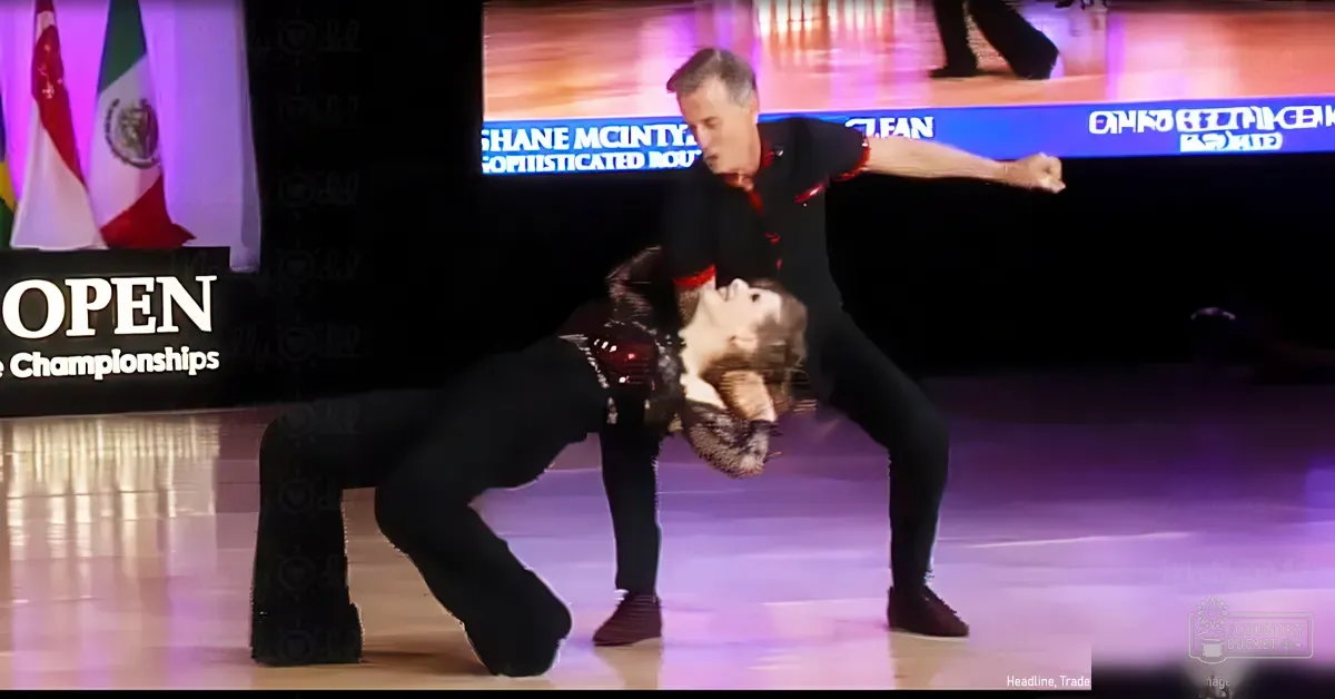 A dance duo performing at an open championship event in a well-lit indoor venue with flags from various countries and a large screen displaying 'OPEN Championships' and names like Shane McIntyre.