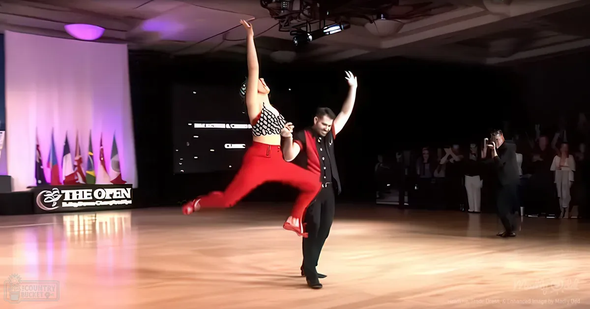 A dance couple performs at 'The Open Ballroom Dance Championship' in an indoor ballroom with wooden flooring and overhead lights, with international flags and spectators.