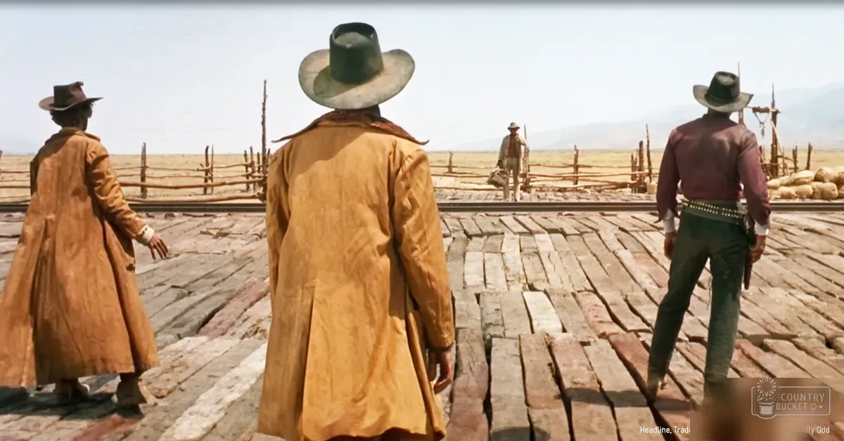 A group of cowboys in long brown coats and wide-brimmed hats stand against an open desert landscape with wooden fences, train tracks, and distant mountains.