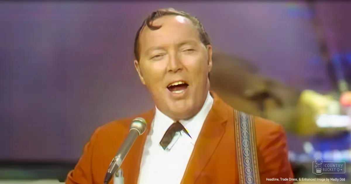 A middle-aged male singer in a vintage orange suit jacket with patterned lapel, white shirt, and dark tie performs on stage.